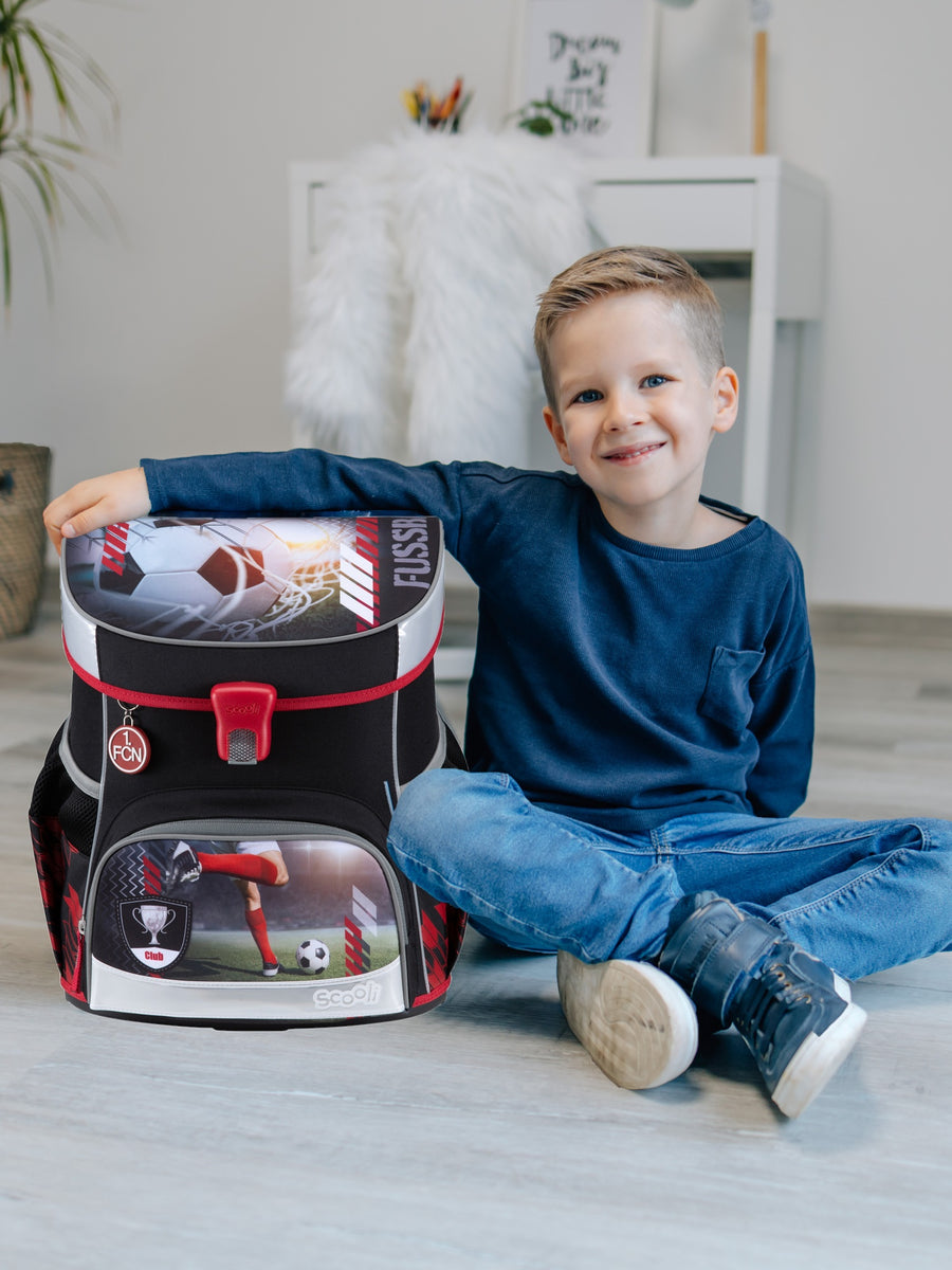 Jungen sitzt lächelnd in hellem Wohnzimmer mit einem Arm um den FCN Schulranzen in schwarz mit roten und grauen Akzenten