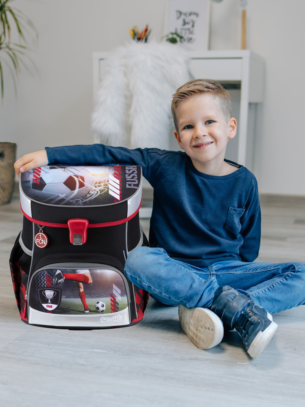 Jungen sitzt lächelnd in hellem Wohnzimmer mit einem Arm um den FCN Schulranzen in schwarz mit roten und grauen Akzenten