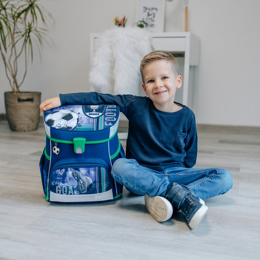 Junge sitzt lächelnd am Wohnzimmerboden und hat Arm auf Fussball Schulranzen gelegt