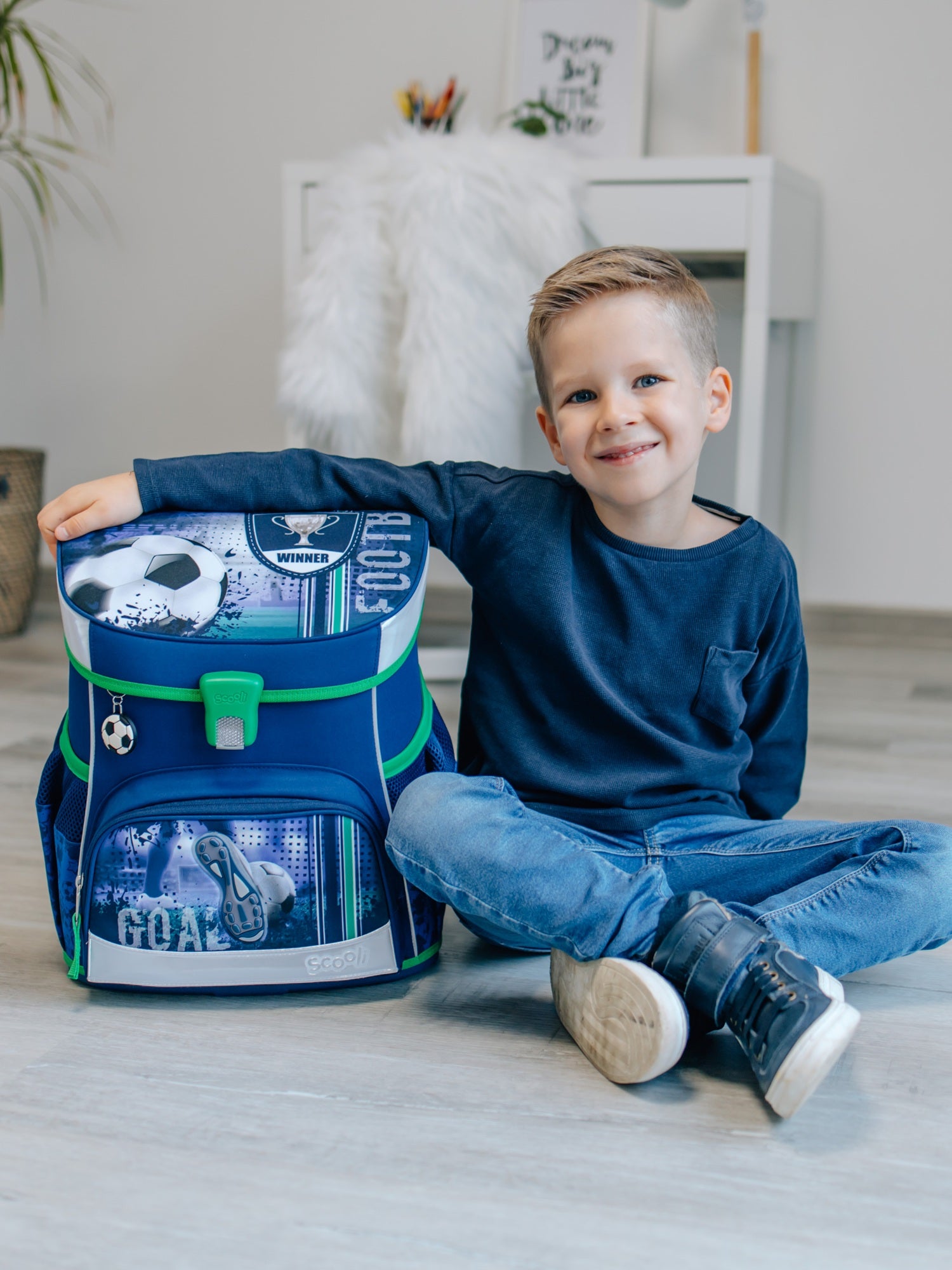 Junge sitzt in hellem Wohnzimmer und lächelt in die Kamerea mit einem Arm um den blauen Fußball Schulranzen