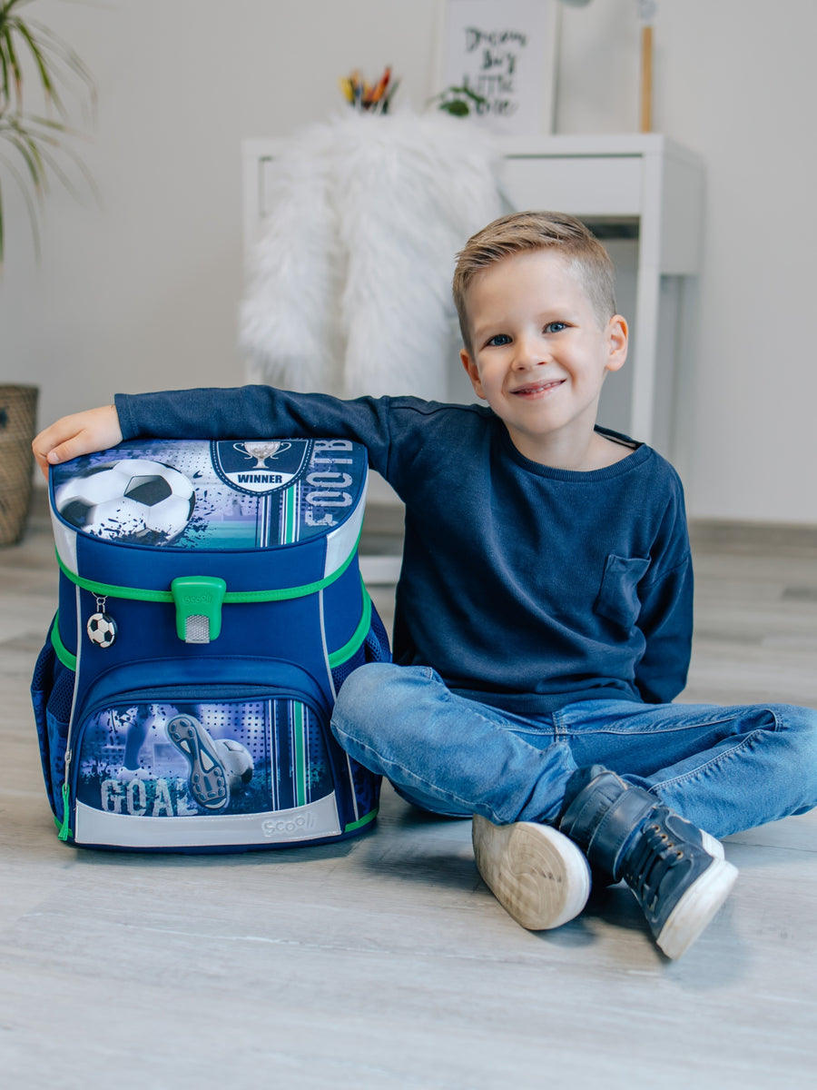 Junge sitzt in hellem Wohnzimmer und lächelt in die Kamerea mit einem Arm um den blauen Fußball Schulranzen