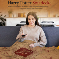 Frau sitzt auf der Couch uns sieht fern während sie sich in die Harry Potter Rumtreiber Decke kuschelt