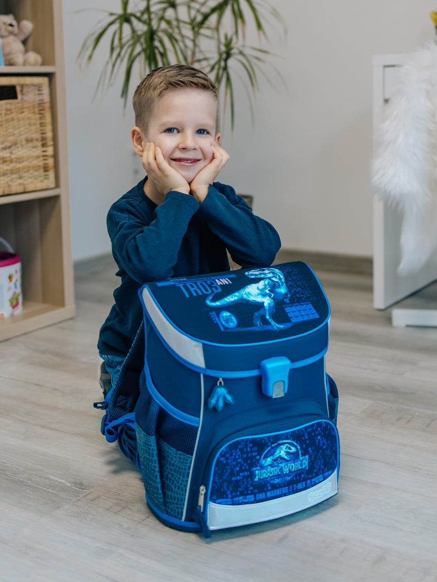Junge kniet in einem hellem Wohnzimmer und lächelt, vor ihm der dunkelblaue Jurassic World Schulranzen