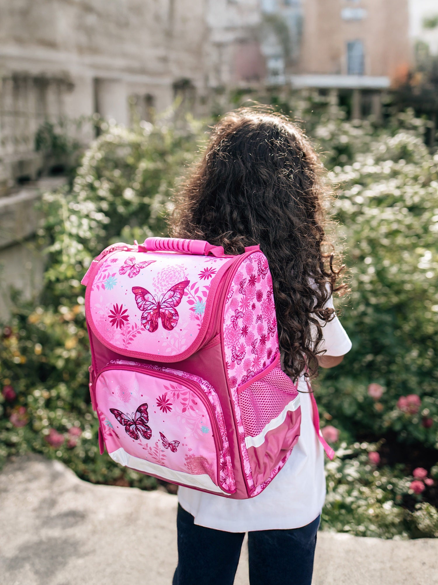 Mädchen mit schwarzen Locken trägt rosa Schmetterlings Schulranzen mit Rücken zur Kamera