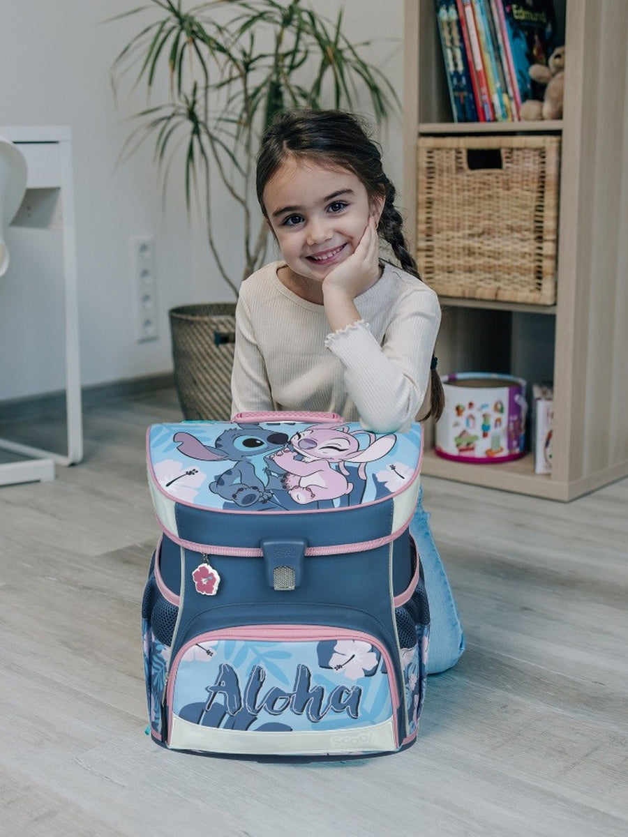 Mädchen kniet in hellem Wohnzimmer mit einem Ellenbogen auf hellblauem Stitch Schulranzen gestützt und lächelt in die Kamera