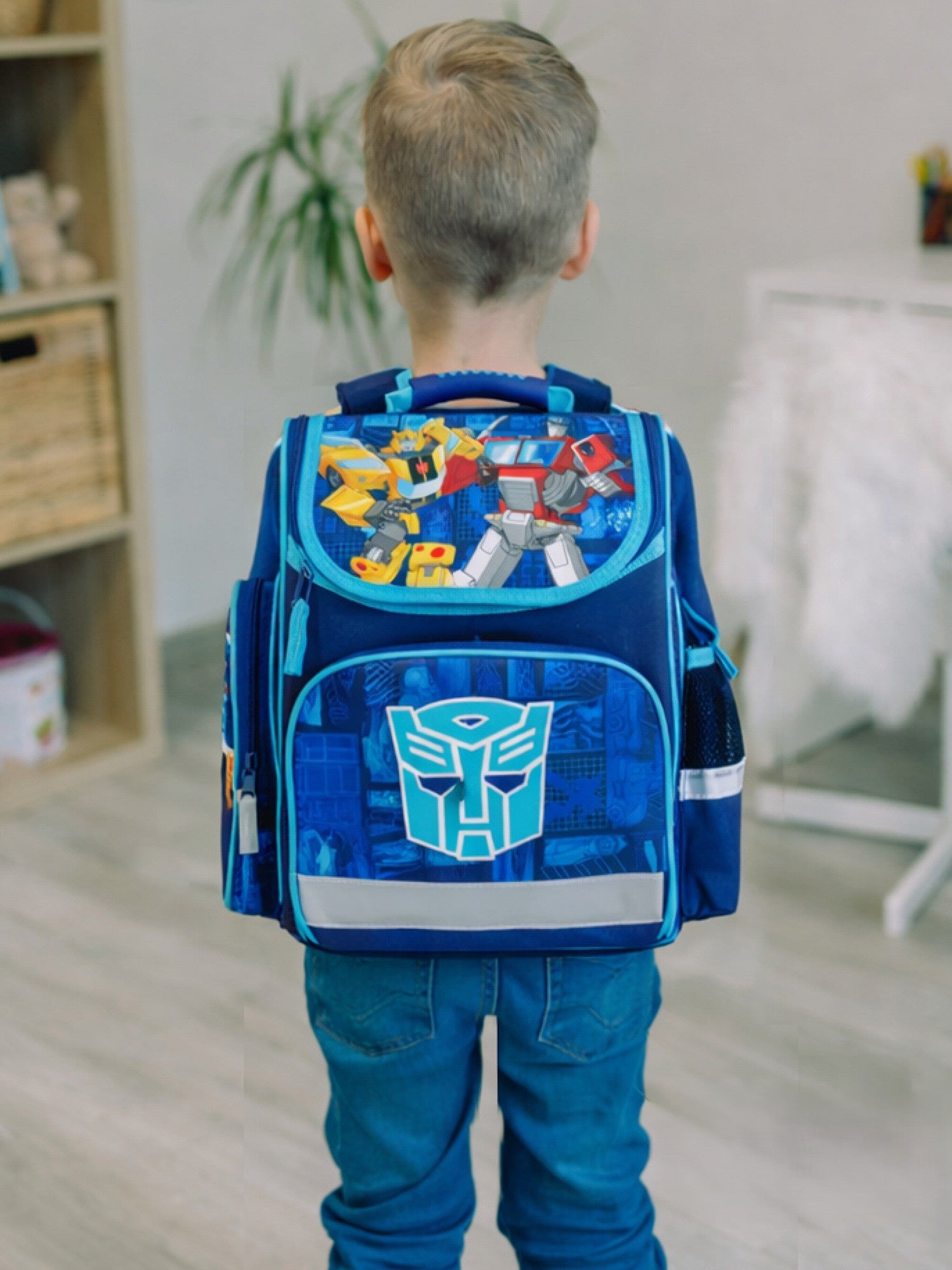 Junge steht mit Rücken zur Kamera und trägt den hellblauen Transformers Schulranzen mit Optimus Prime und Bumblebee auf Deckel am Rücken